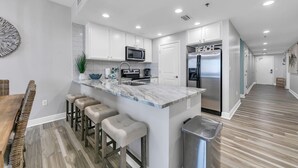 A modern kitchen with white cabinets, a marble countertop, and stainless steel appliances. There are three bar stools and a trash can.