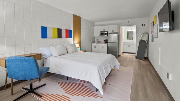 A modern hotel room with a bed, a desk, a chair, a kitchen area, and a TV mounted on the wall. - Stay Downtown, Free Gated Parking, Renovated Studio! 512 (Cedar Falls)