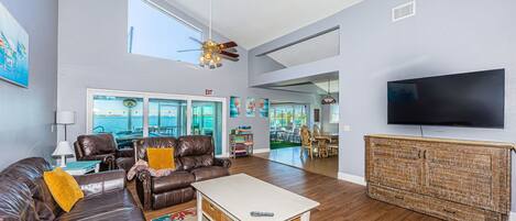 A spacious living room with a leather sofa, a white coffee table, and a television. There is a large window offering a view of the water and a balcony.