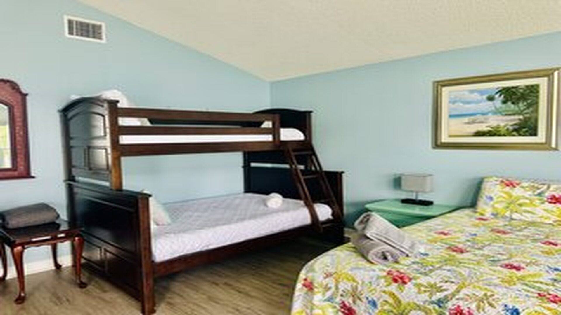 A room with a bunk bed, a desk, a chair, a nightstand with a lamp, and a bed with a floral bedspread.