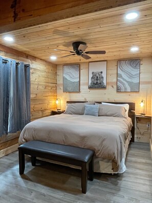 A wooden-paneled bedroom with a bed, bench, ceiling fan, and wall art. - Walk to Midtown, King Bed, Cozy Cabin! (Ruidoso)