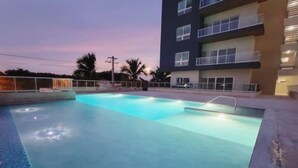 An outdoor swimming pool with a clear blue water surface, surrounded by a low concrete wall and a metal railing.