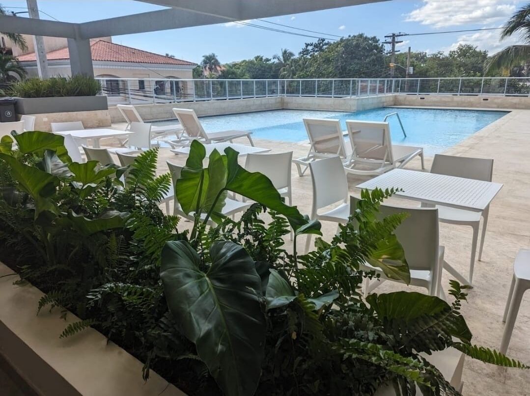 A poolside area with white lounge chairs and tables, surrounded by lush green plants.