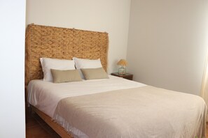 Een bed met een geweven hoofdbord, witte lakens en een klein nachtkastje met een lamp.
