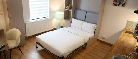 A compact bedroom with a bed, a chair, a desk, a bedside table, a lamp, and a small kitchenette.