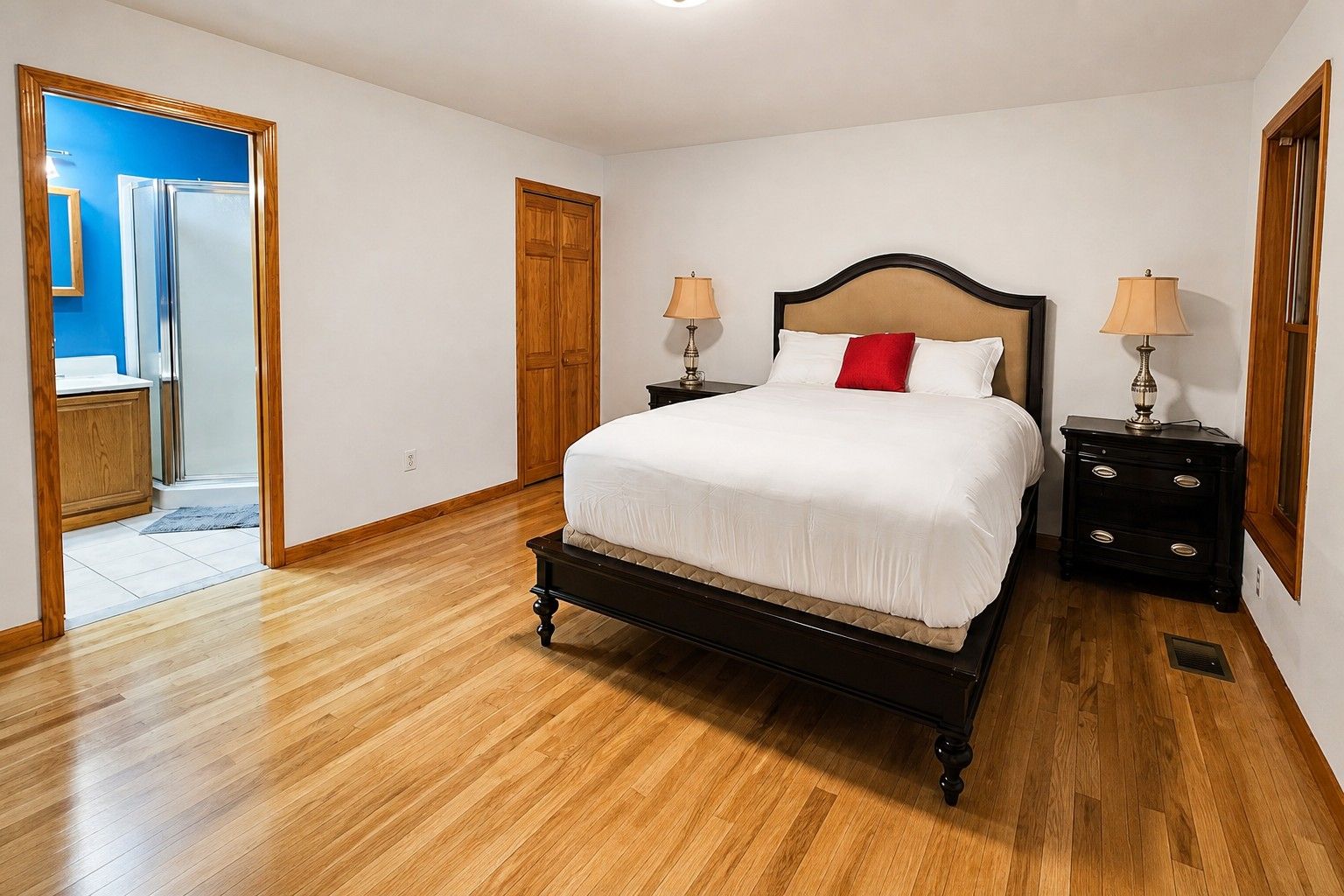 A bedroom with a wooden floor, a bed with white bedding and a red pillow, two bedside tables with lamps, and a wooden door.