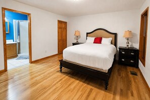 Une chambre avec un plancher en bois, un lit avec une literie blanche et un oreiller rouge, deux tables de chevet avec des lampes et une porte en bois.