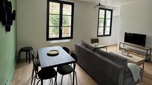 Ein modernes Wohnzimmer mit Essbereich, einer Couch, einem Fernseher und einem Fenster mit Blick auf Grünflächen.