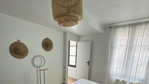 Ein Schlafzimmer mit einem großen Bett, einer hängenden Lampenschirm aus Rattan, einem runden Spiegel und Blick ins Freie durch ein Fenster.