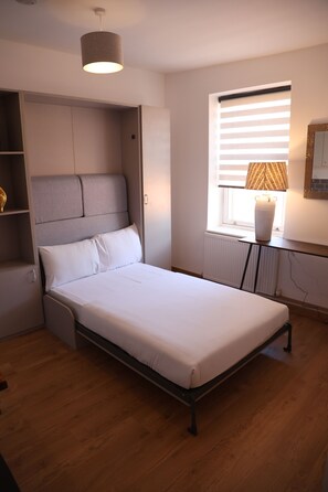 Un dormitorio con cama, almacenamiento integrado, ventana con persianas y lámpara en una mesita de noche.