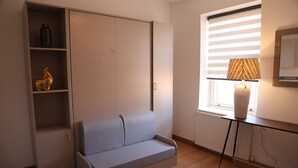 A room with a grey sofa, a wardrobe with shelves, a radiator, and a window with blinds. - Stylish Studio Escape (London)