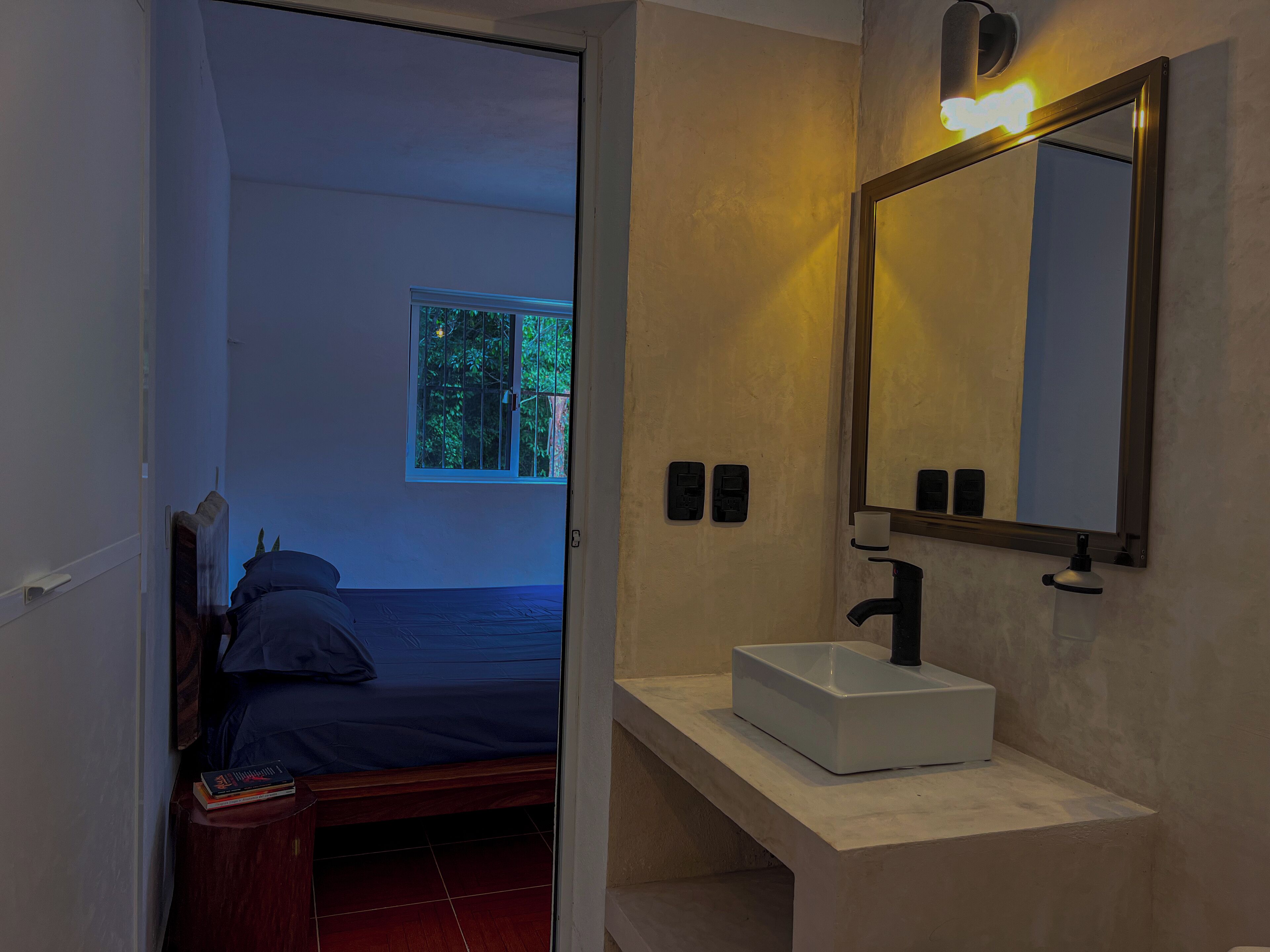A bedroom with a bed, a bedside table with a book, a window with a view of greenery, and a bathroom with a sink and mirror.