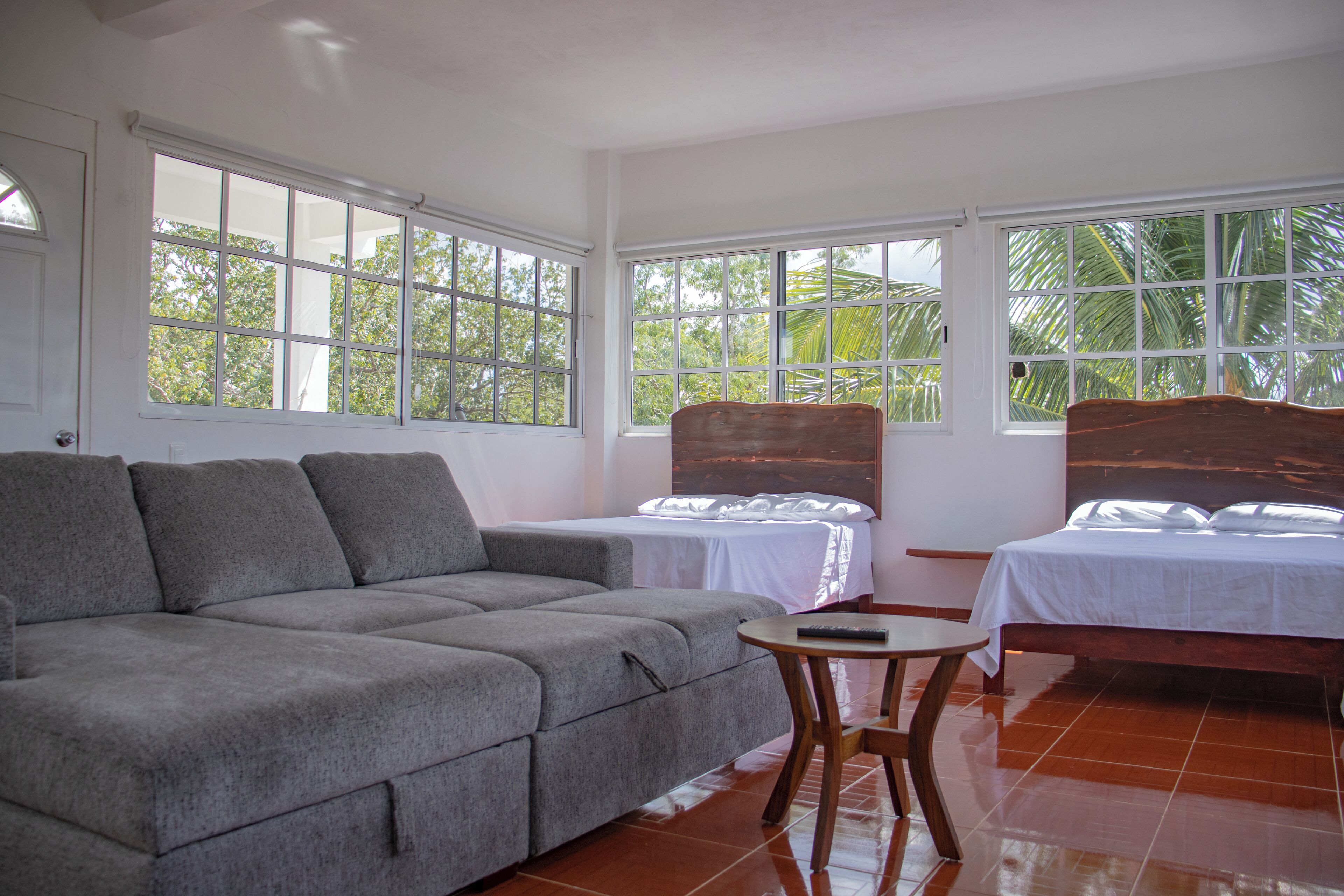 Ein Zimmer mit grauem Sofa, zwei Einzelbetten, einem kleinen Tisch und großen Fenstern mit Blick auf Palmen.