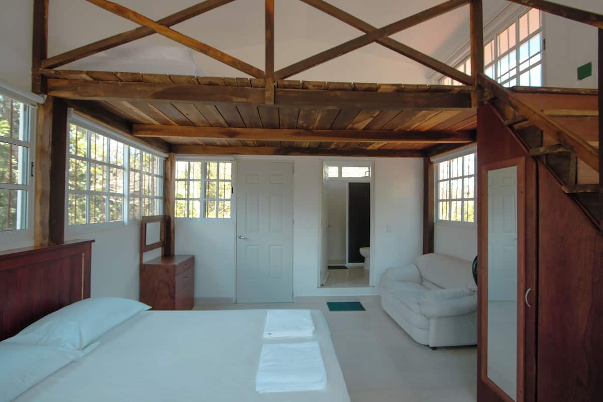 Een slaapkamer met een houten plafond, een bed, een dressoir, een stoel en een deur naar een badkamer.