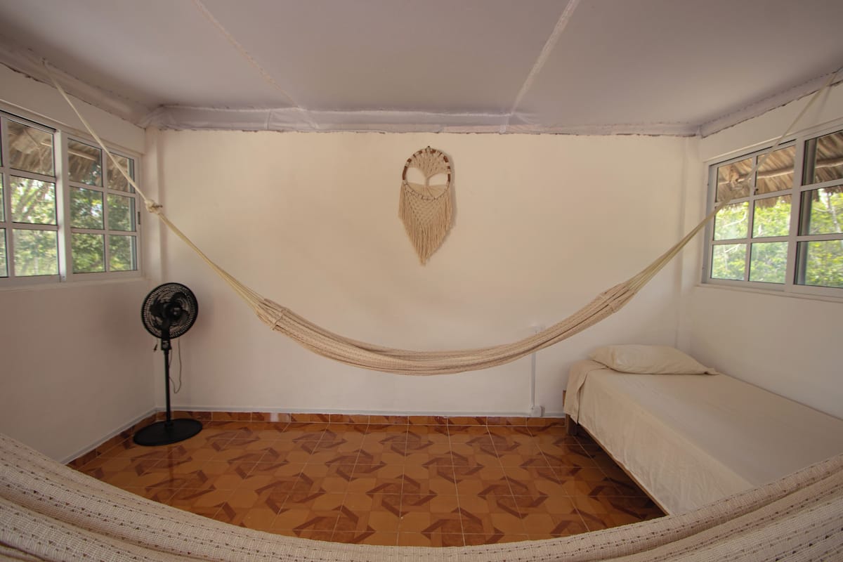 Een kamer met een hangmat, een bed, een staande ventilator en vloertegels met een patroon.