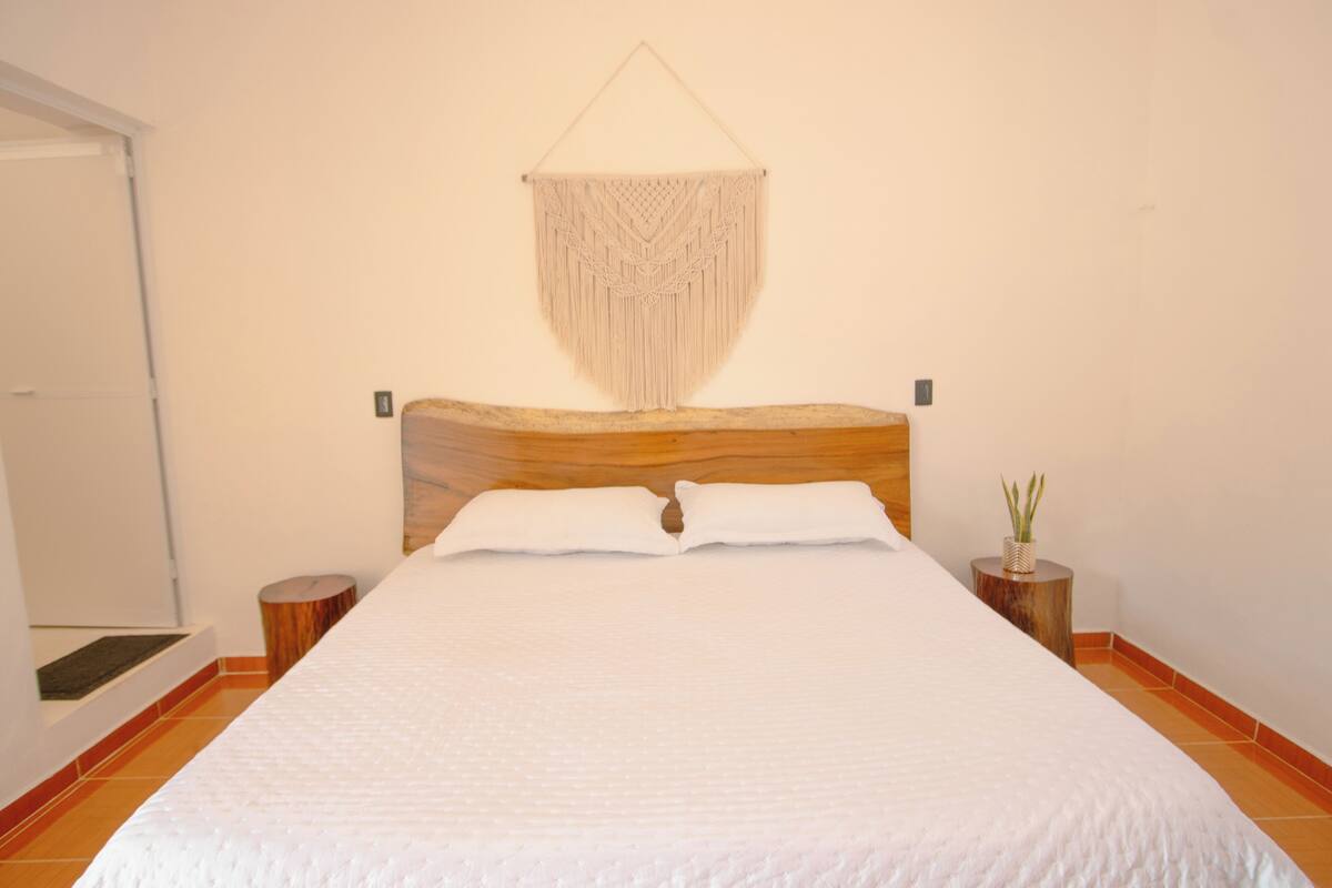 Een net opgemaakt bed met witte lakens, een houten hoofdbord en een macramé wanddecoratie.