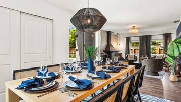 A dining area with a long wooden table set for six, surrounded by chairs with woven backs. A large hanging light fixture is above the table.