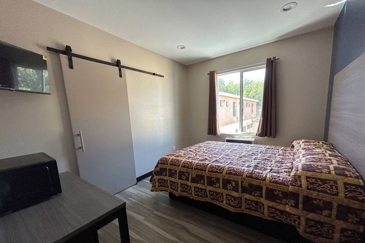 A bedroom with a bed, a desk, a window with curtains, and a view of buildings outside.