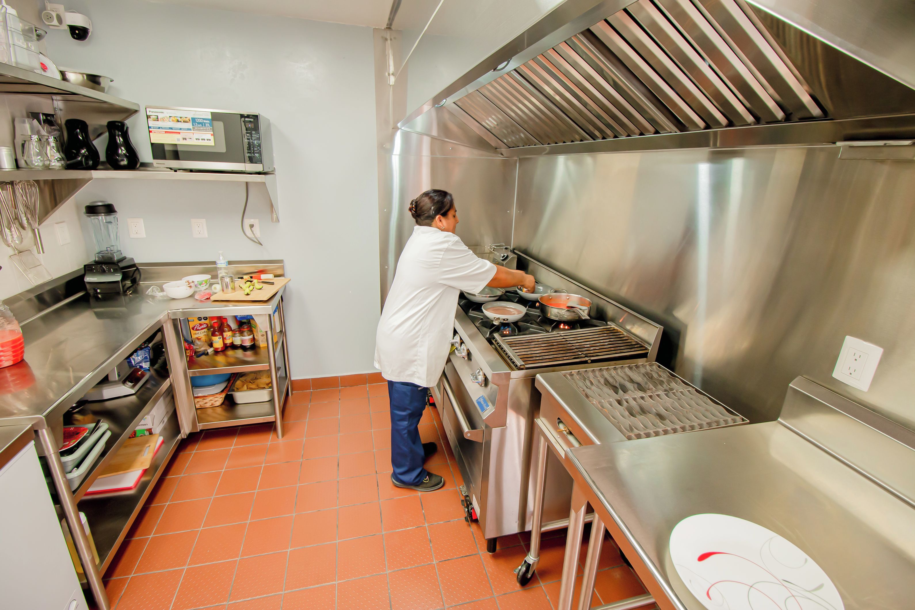 A commercial kitchen with stainless steel appliances, a person cooking, and various kitchen tools and ingredients.