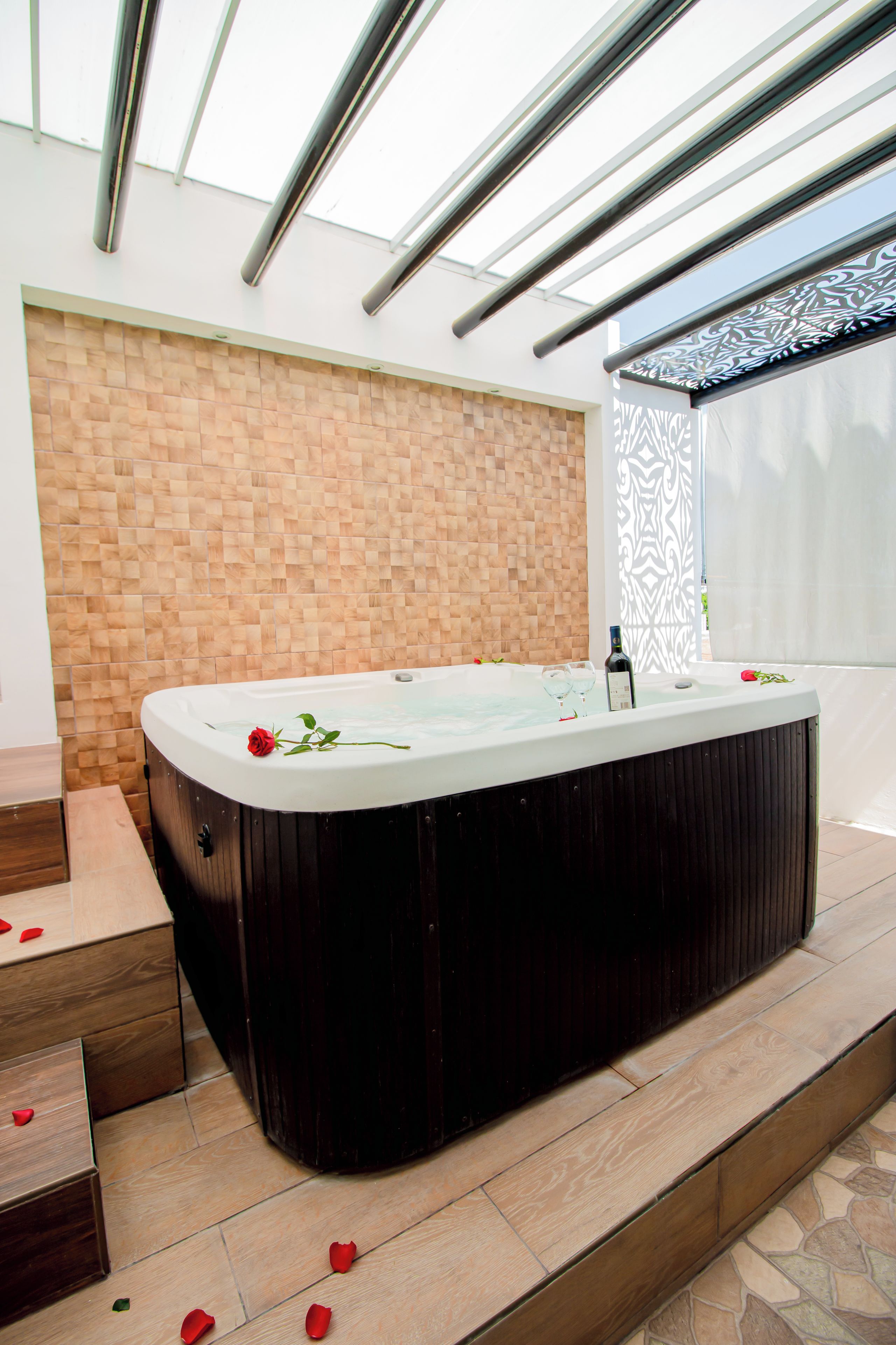 A hot tub with rose petals on the floor, a wooden wall, and a window with a patterned blind.