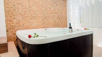 A hot tub with rose petals on the floor, a wooden wall, and a window with a patterned blind.