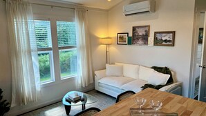Una habitación con un sofá blanco, una mesa de centro de vidrio, una mesa de comedor de madera con copas de vino, y vistas a zonas verdes a través del ventanal.