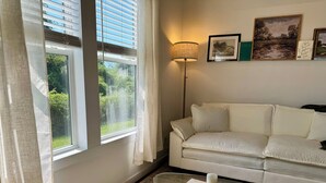 Un salón con un sofá blanco, una mesa de centro de vidrio con un libro y una vela, una lámpara de pie y vistas a la vegetación por la ventana.