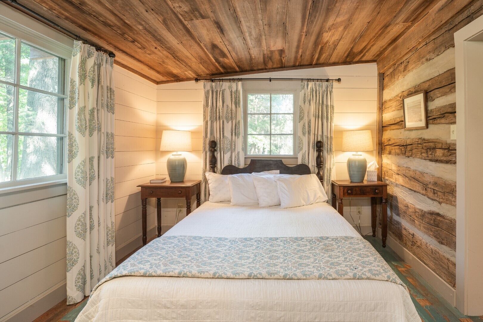A cozy bedroom with a wooden ceiling, two bedside tables with lamps, a bed with white bedding, and a window with curtains.