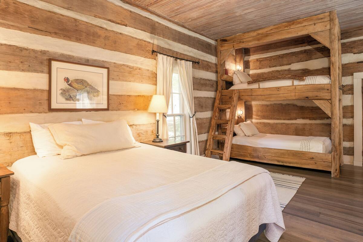 Ein schlicht eingerichtetes Schlafzimmer im Chalet-Stil mit einem großen Bett, einem Schlafplatz in der Schlaffküche, Holzwänden, einem Bilderrahmen mit einer Vogelansicht und einem Nachttisch mit Lampe.