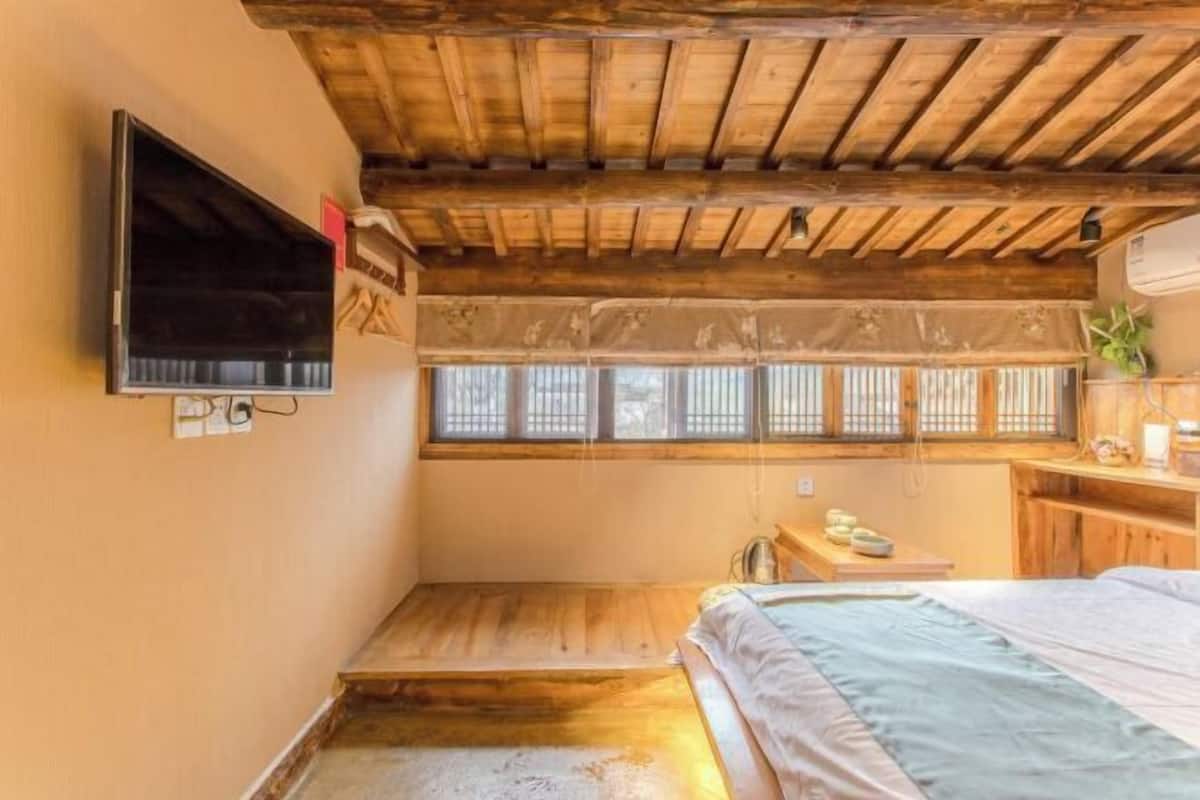 A small, cozy room with a wooden ceiling, a bed, a small table, and a TV mounted on the wall.