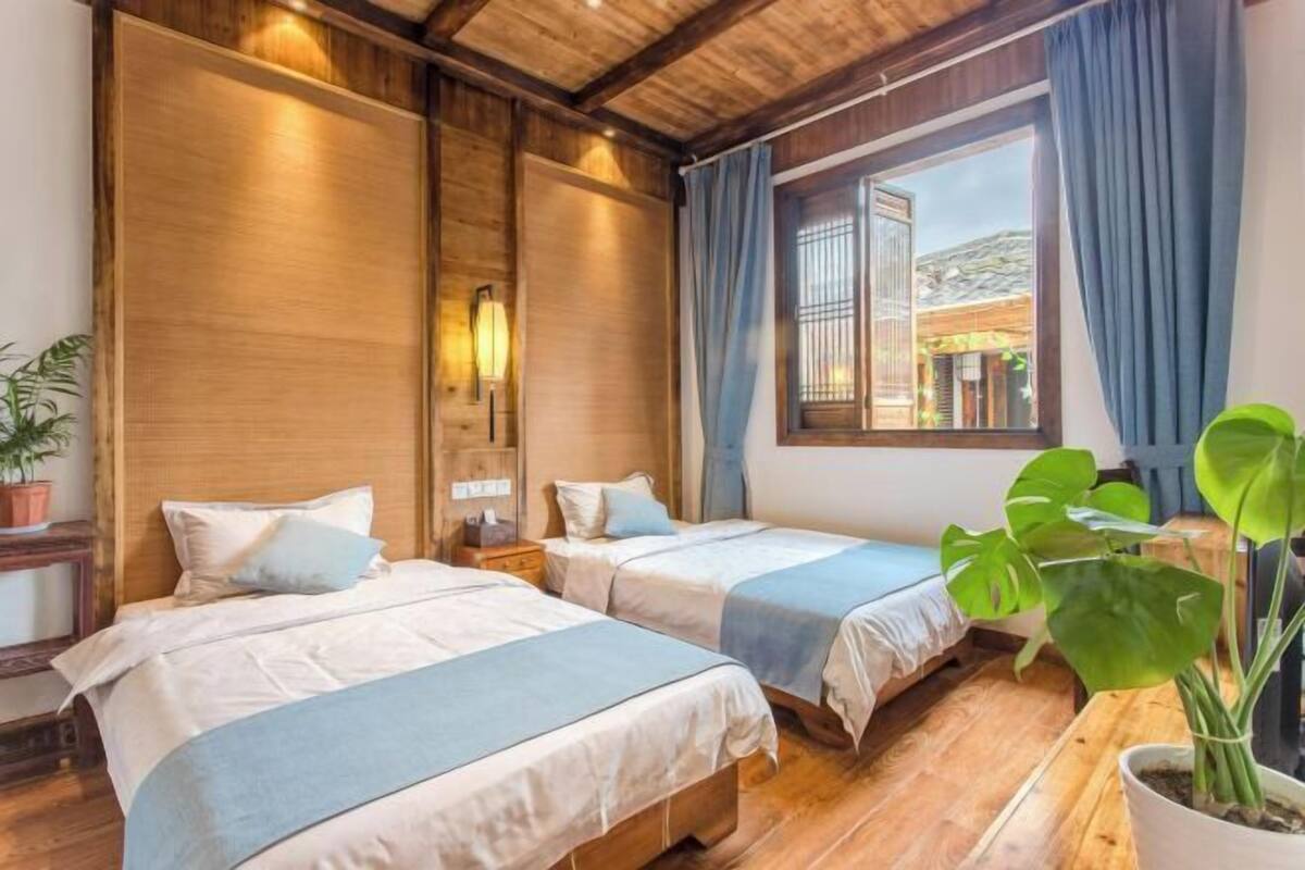 A room with two beds, wooden walls, a large window with curtains, and a potted plant.