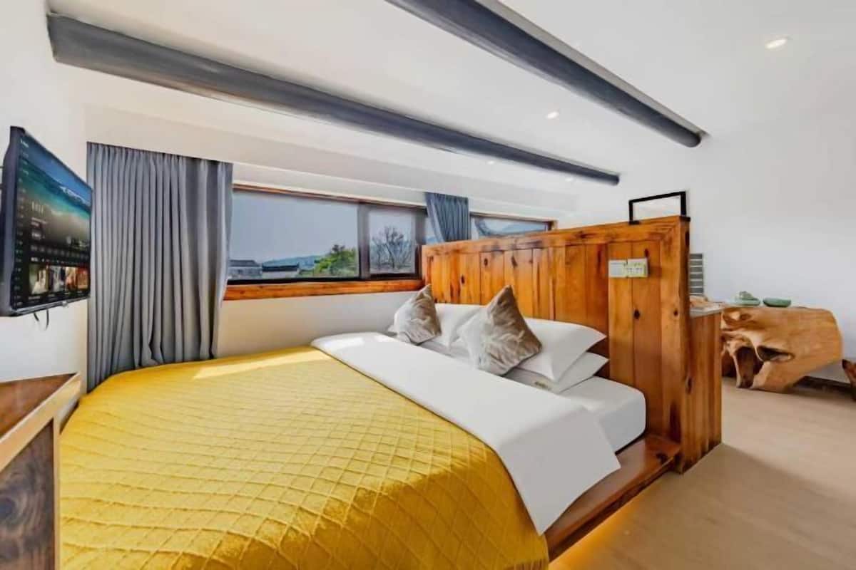 A hotel room with a bed, a wooden headboard, a television, a window with curtains, and a view of the outside.