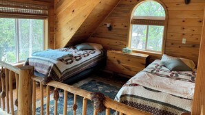 A cozy attic bedroom with two beds, wooden walls, a window, and a small table.
