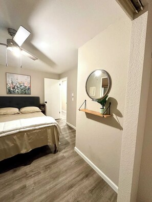 A bedroom with a bed, a ceiling fan, a wall-mounted mirror, and a small shelf with a plant.