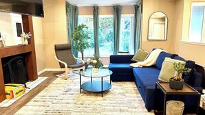 Un salon avec un canapé bleu, une table basse en bois, un fauteuil, une cheminée et une grande fenêtre avec des rideaux.