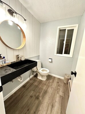 A bathroom with a wooden vanity, a round mirror, a toilet, and a window.