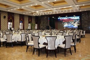 Un salón de banquetes con mesas y sillas redondas, un escenario con una pantalla y una araña.