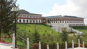 A large building with a sloped roof, situated on a hill with a paved walkway and a fence in the foreground.