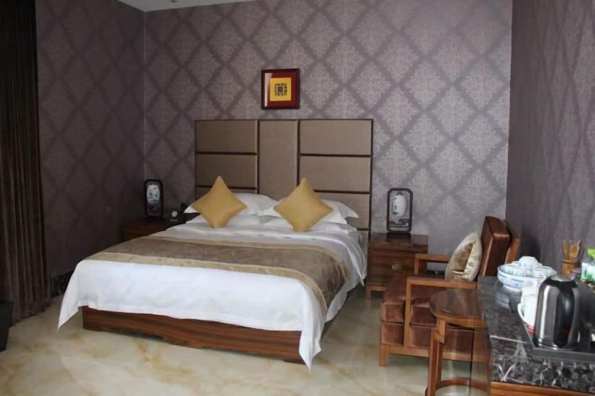 A hotel room with a large bed, wooden headboard, two bedside tables with lamps, a wooden desk with a chair, and a framed picture on the wall.