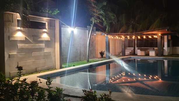 Un espace piscine nocturne au design moderne, comprenant une piscine entourée de parois en verre et un patio illuminé de guirlandes lumineuses.