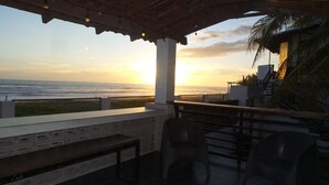 Un espace extérieur aménagé avec des sièges, offrant une vue sur l’océan au coucher du soleil, et comprenant une pergola en bois, des murs en pierre et des fauteuils en osier.