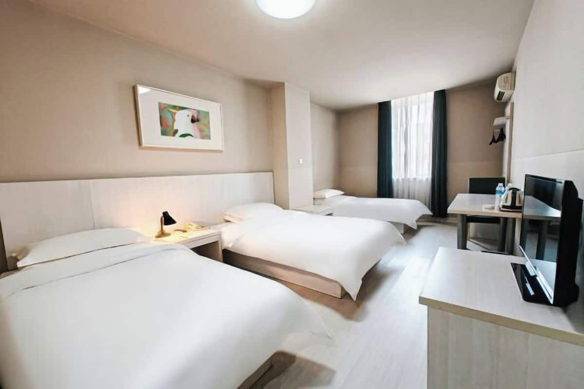 Habitación de hotel con tres camas, un escritorio, un televisor y aire acondicionado instalado en la pared.