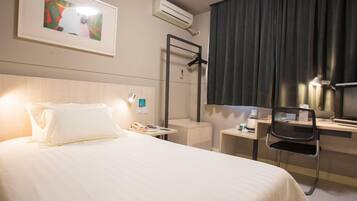A hotel room with a bed, bedside table, chair, desk, and a painting on the wall.