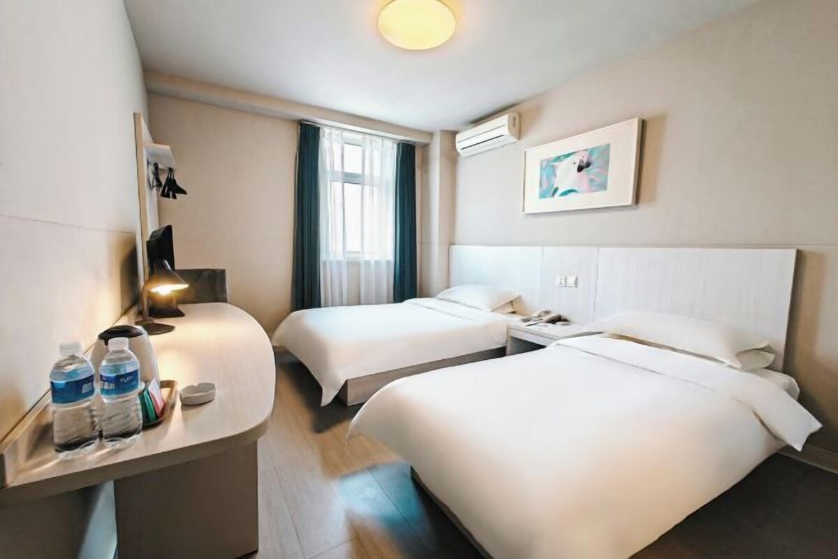 A hotel room with two beds, a desk with water bottles, a bedside table, a window with curtains, and a wall-mounted air conditioner.