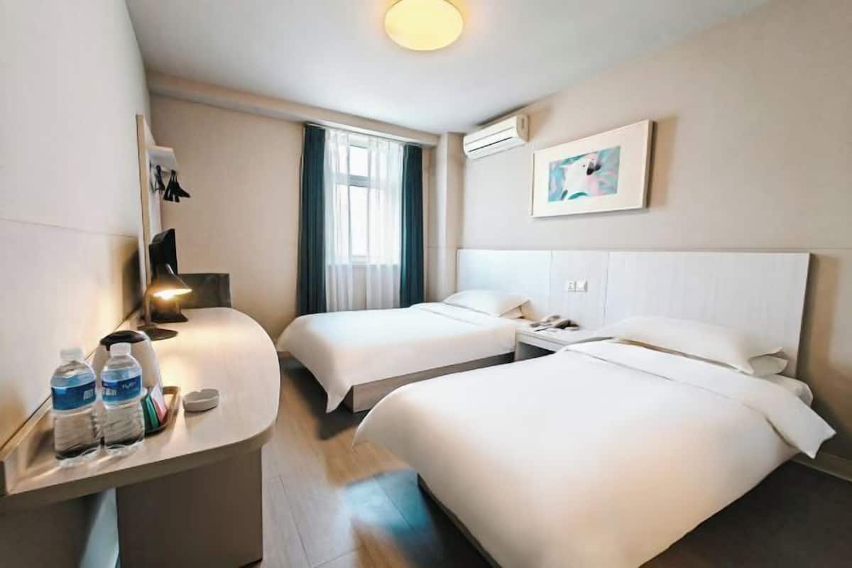 Habitación de hotel con dos camas, un escritorio con botellas de agua, una mesita de noche, una ventana con cortinas y un aire acondicionado instalado en la pared.