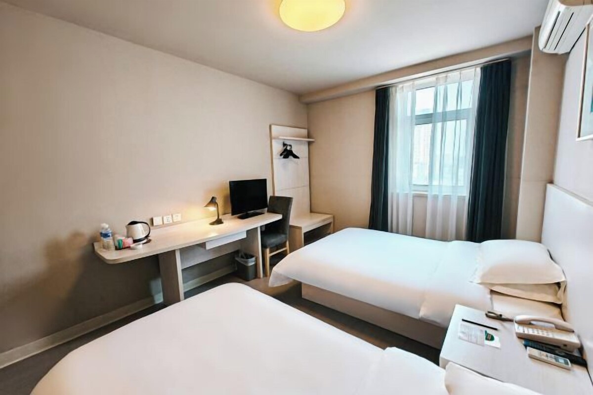 A hotel room with two beds, a desk with a computer, a TV, and a bedside table with a phone and remote controls.