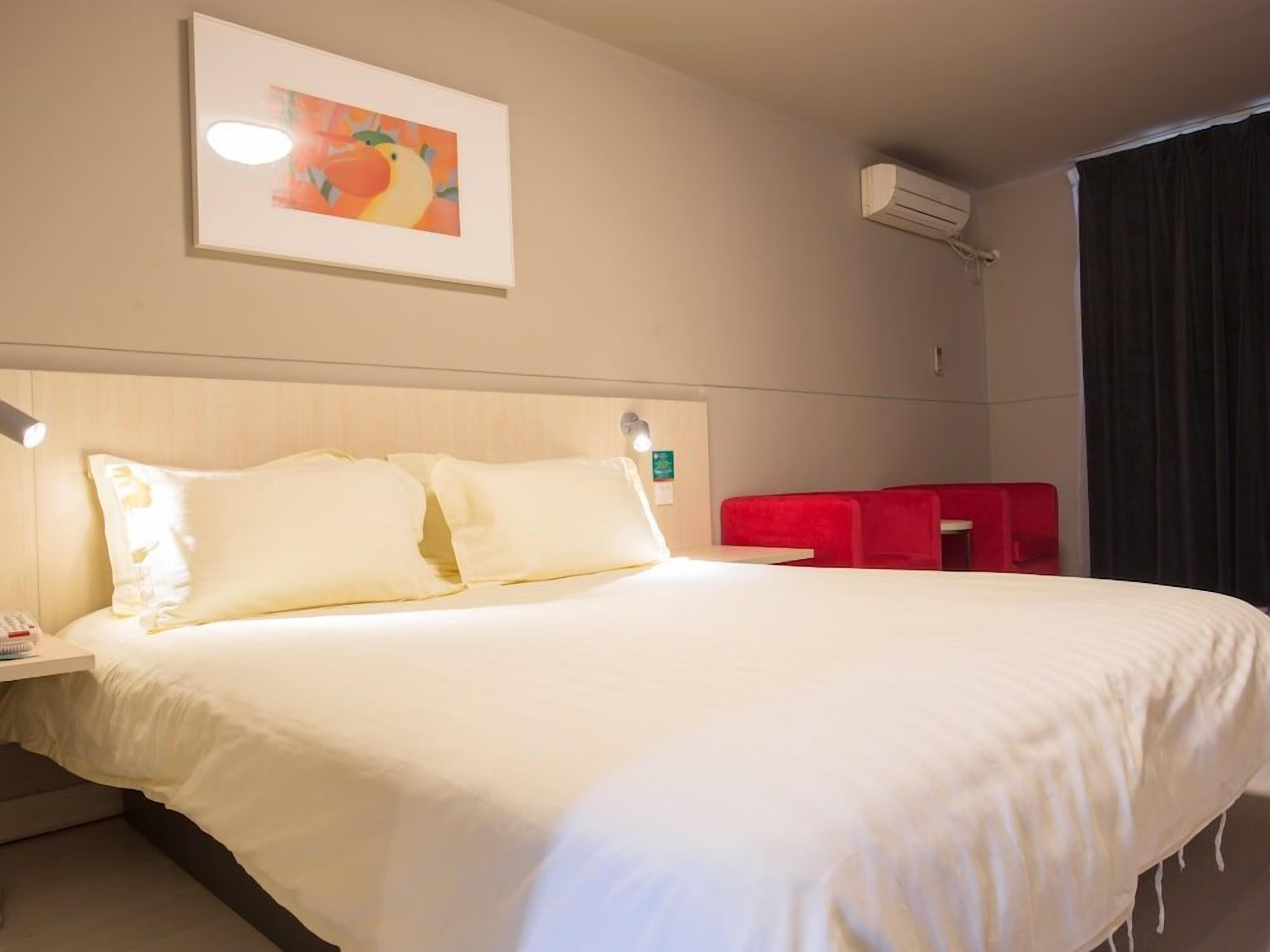A hotel room with a large bed, a nightstand, a red chair, a wall-mounted air conditioner, and a framed picture above the bed.