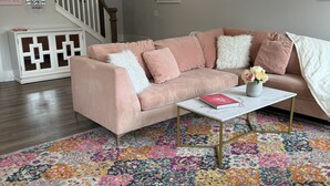 Una sala de estar con un sofá seccional rosa, una mesa de centro blanca y una alfombra colorida.