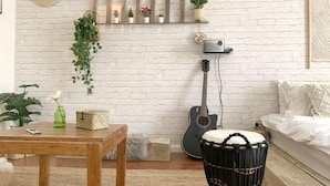 Een knus woongedeelte met een witte bakstenen muur, een houten salontafel, een gitaar, een drumstel en een plank met planten.