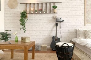 A cozy living room with a white brick wall, a wooden coffee table, a guitar, a drum, and a shelf with plants.